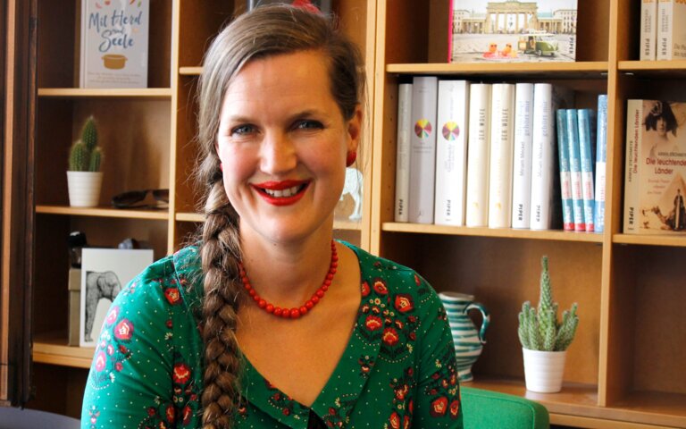 Frau in grünem Shirt blickt unmittelbar und lächelnd in die Kamera. Im Hintergrund steht ein Bücherregal mit vorwiegend weißen Buchrücken. | © Foto: Piper Verlag