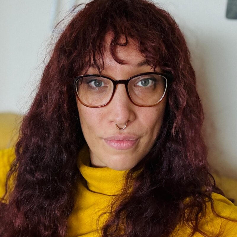 Das Foto zeigt Jasmin Dickerson. Sie hat lange rotbraune Haare, trägt eine Brille, einen gelben Pullover und sitzt auf einer gelben Couch. | © Die Autorin Jasmin Dickerson wünscht sich mehr Bewusstsein für Neurodiversität. Foto: privat