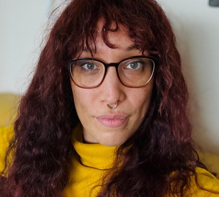 Das Foto zeigt Jasmin Dickerson. Sie hat lange rotbraune Haare, trägt eine Brille, einen gelben Pullover und sitzt auf einer gelben Couch. | © Die Autorin Jasmin Dickerson wünscht sich mehr Bewusstsein für Neurodiversität. Foto: privat