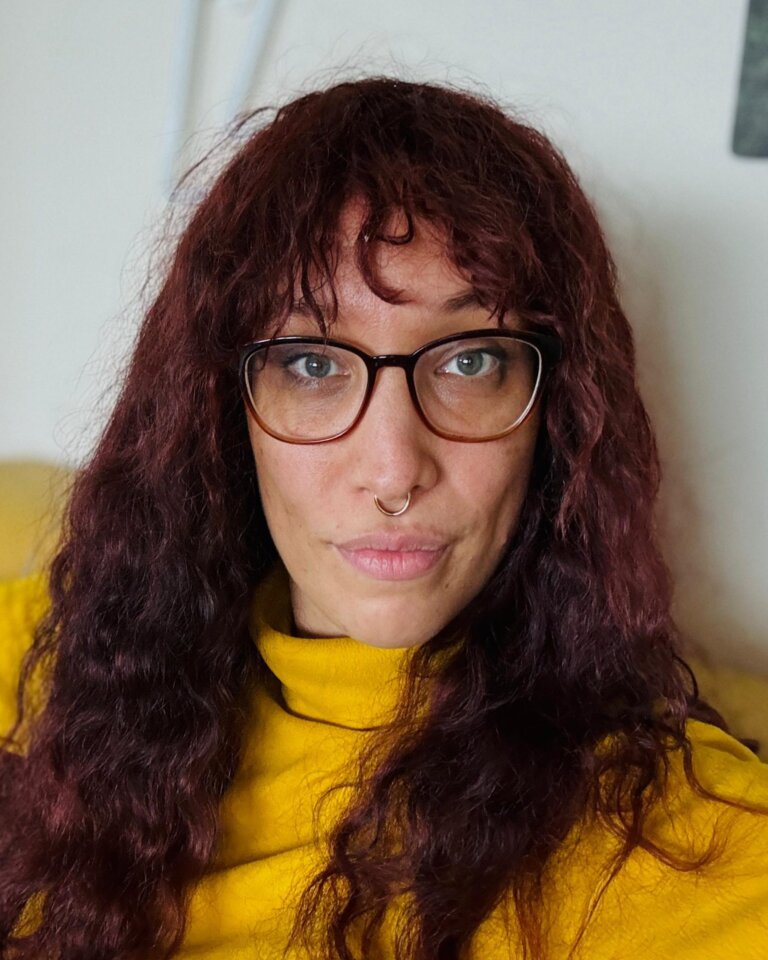 Das Foto zeigt Jasmin Dickerson. Sie hat lange rotbraune Haare, trägt eine Brille, einen gelben Pullover und sitzt auf einer gelben Couch. | © Die Autorin Jasmin Dickerson wünscht sich mehr Bewusstsein für Neurodiversität. Foto: privat